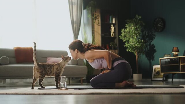 A woman practices yoga on a mat in her living room. A cat walks around her and plays near a water glass. The room has plants and bright natural light coming through the window.