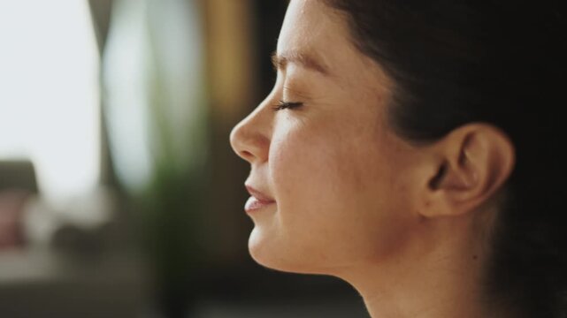A woman sits with her eyes closed, focusing on her breath. She is in a quiet indoor space, taking a moment for herself to relax and clear her mind. The atmosphere looks calm.