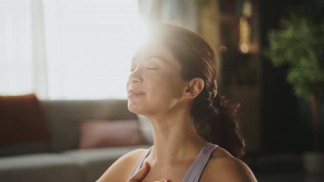 A woman practices a breathing exercise at home in her living room. She sits comfortably and focuses on her breath while keeping her hand on her chest.
