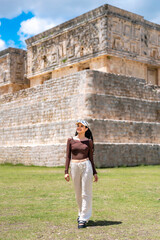 Fototapeta premium Young woman walks forward smiling calmly and confidently while exploring ancient stone structures in Uxmal Yucatan Mexico, enjoying cultural travel, wearing cap, brown top and light loose pants