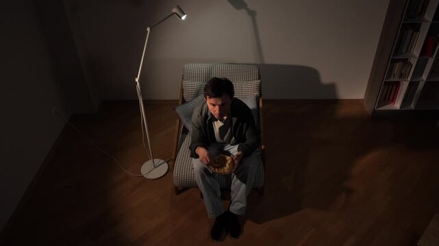 Lonely man sitting on a chair, eating potato chips from a bowl under the light of a floor lamp in a dim room, representing solitude and comfort eating