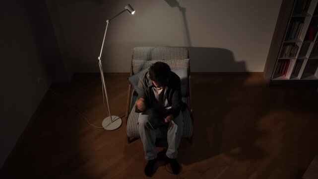 Young man sitting in an armchair, concentrating on a video game, holding a controller in a dim room with a floor lamp providing focused light, highlighting nocturnal entertainment