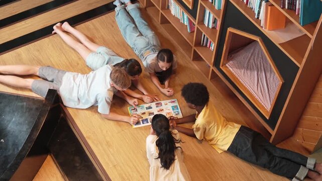 Group of diverse children lying down in circle while reading a book at library. Top view of girl sharing a magazine while pointing at interested topic and talking with lovely friends. Edification