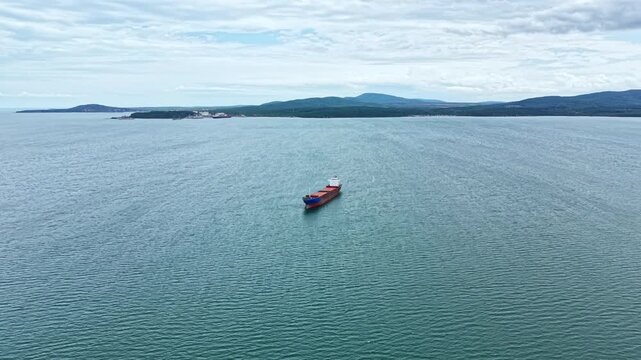 A cargo ship is located in the water with the coastline and hills visible in the background. The sky is filled with clouds during the autumn season.