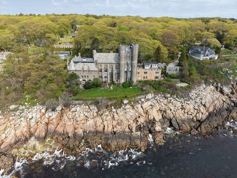 Stunning aerial view of Hammond Castle on the rocky coast of Gloucester, Massachusetts. This historic medieval-style estate sits on a cliffside overlooking the ocean, surrounded by lush spring trees.