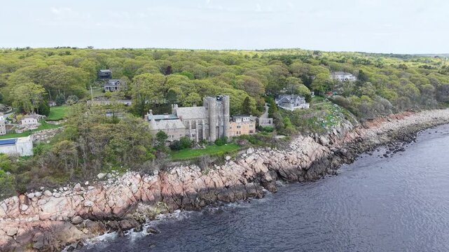 Stunning aerial view of Hammond Castle on the rocky coast of Gloucester, Massachusetts. This historic medieval-style estate sits on a cliffside overlooking the ocean, surrounded by lush spring trees.