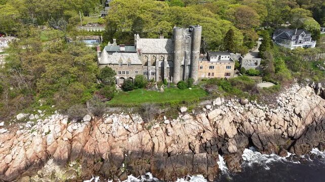 Stunning aerial view of Hammond Castle on the rocky coast of Gloucester, Massachusetts. This historic medieval-style estate sits on a cliffside overlooking the ocean, surrounded by lush spring trees.