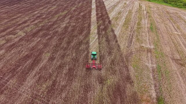 A large tractor is plowing a wide farm field. The ground shows rows of tilled soil. The surrounding area has green plants and trees. The sun is shining in the sky.