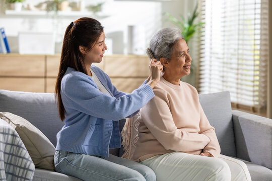 Young woman caring for elderly mother adjusting hearing aid device on couch at home, expressing warmth, love, compassion, and family connection in comfortable bright living room environment