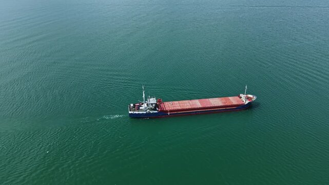 A cargo ship travels on bright blue water. The sea is smooth and stretches far into the distance. The sun shines on the ship while it carries its load.