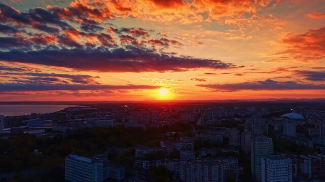 A bright sunset occurs over a city with tall buildings. The sky has clouds and many colors. People enjoy the view from a high location in the evening time.