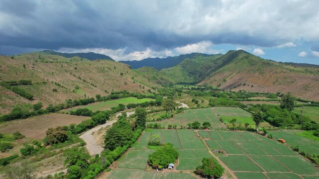 Aerial drone footage of a long dry river in Kilo city area, Bima regency, Sumbawa island, Indonesia, with red onion fields, agricultural areas, and green hills behind, in a dark cloudy weather