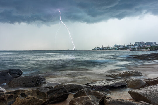 Storm clouds and lightning strike over ocean with rocky shoreline and coastal town in background