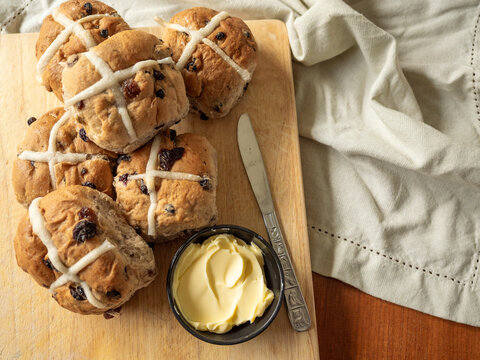 Half dozen Hot Cross Buns on wooden cutting board with butter and knife and linen napkin