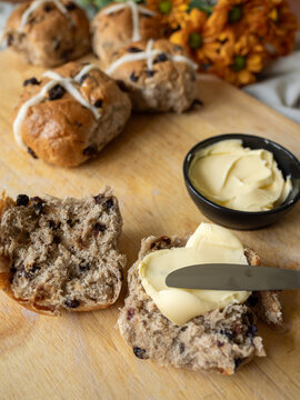 Close up of cut Hot Cross Bun with butter and knife on cutting board with full buns and flowers