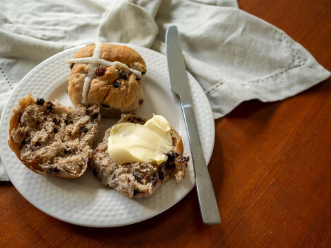 Hot cross buns with butter and knife on white plate, linen napkin and wooden tabletop