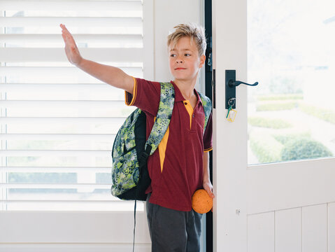 Young boy wearing backpack waving goodbye at the doorway