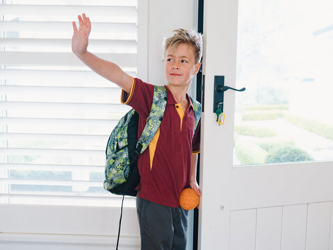 Young boy wearing backpack holding a small orange ball waving goodbye at the doorway