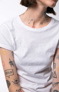 Tiny patchwork tattoos across arms and collarbone with hearts, arrows, and abstract symbols on person wearing white t shirt