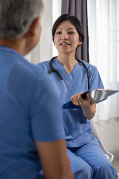 Asian female nurse talking with senior man patient holding clipboard expressing empathy care compassion helpfulness inside medical clinic room Vertical