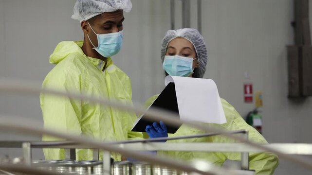 Supervisory team wearing PPE meticulously inspects the mackerel or sardine production line from canning to tomato sauce filling on conveyor belts in industrial seafood processing plants.