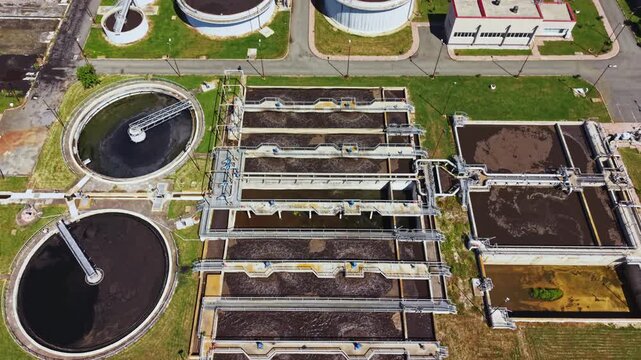 Aerial wastewater treatment plant view shows circular clarifiers, rectangular aeration basins, storage tanks, piping networks, sludge layers, concrete walkways,
