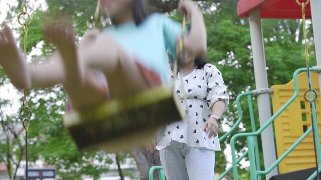 Caring Asian mother pushing her young daughter on a swing at the outdoor playground.
