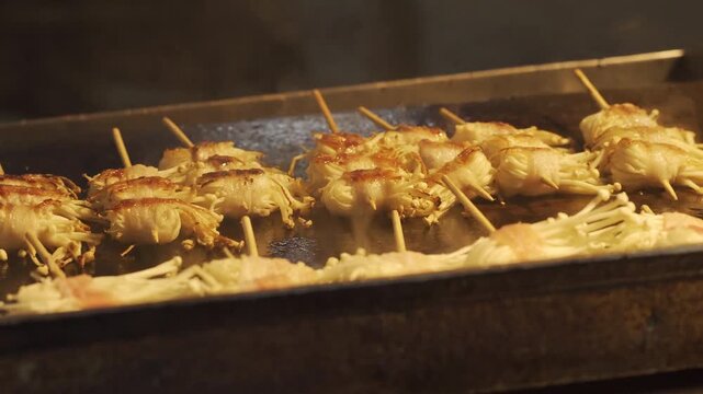 Close-up of bacon wrapped enoki mushroom skewers cooking on a flat top grill at a street food market.