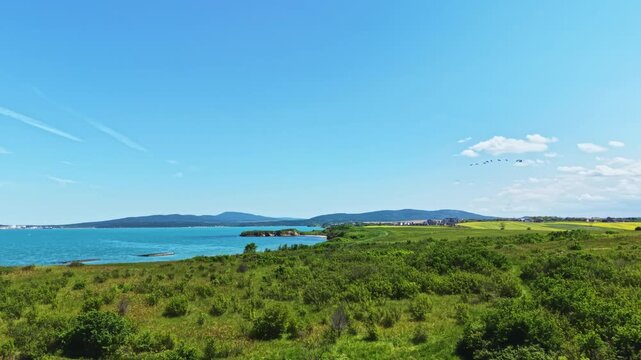 Coastal shrubland overlooking turquoise ocean inlet, wide panoramic view revealing calm azure sea, distant islands on horizon, verdant low shrubs, sandy pebble
