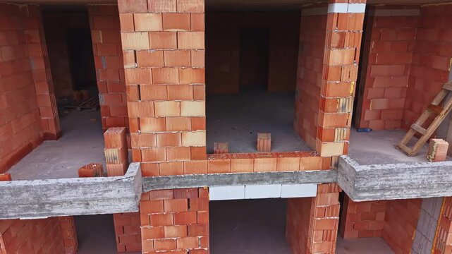 Unfinished brick building with empty openings. Raw red brick walls, exposed concrete slabs, scattered debris, wooden pallets, mortar buckets, void apartment