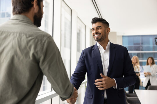 Two cheerful multiethnic businessmen greeting each other with handshake gesture while standing in modern conference room of corporate office, start or accomplish negotiation. Trust and collaboration