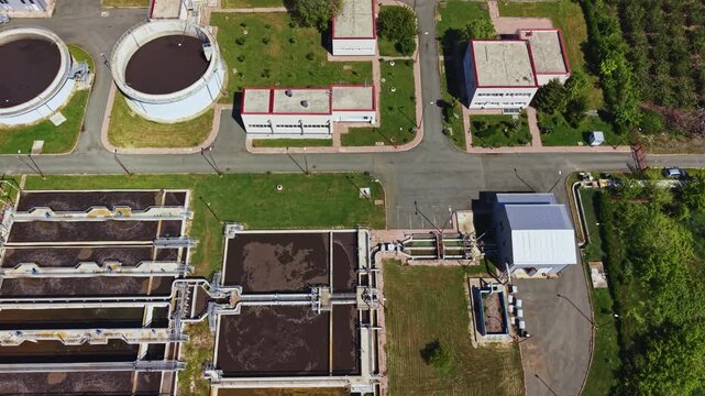 Aerial sewage treatment plant showing clarifiers and rectangular aeration basins, circular settling tanks, access roads, maintenance shed, green buffer
