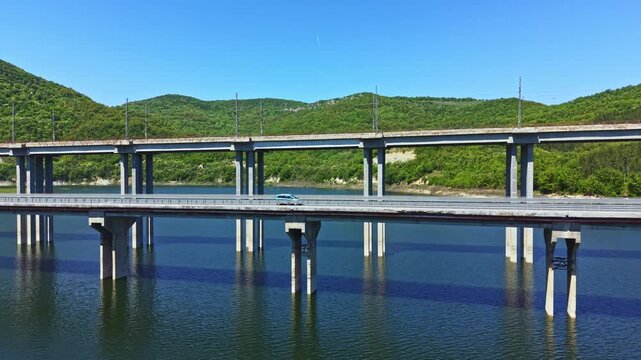 Cars move along a road on the lower section of a bridge that spans over a river. The upper bridge supports train tracks. Lush green hills surround the area. It is a bright sunny day.