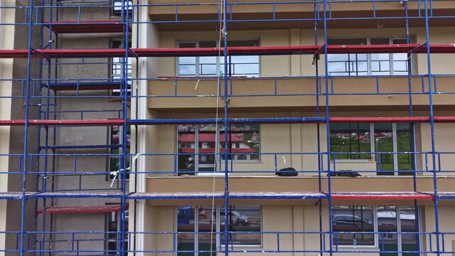 Scaffolding covered apartment facade mid renovation, metal frames and platforms span multiple floors, beige cladding and window installations visible,