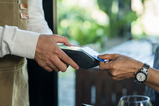 Asian son presents payment terminal to customer in family cafe, practicing cashless checkout, card transaction, front counter service, restaurant billing workflow, mentorship through daily operation.