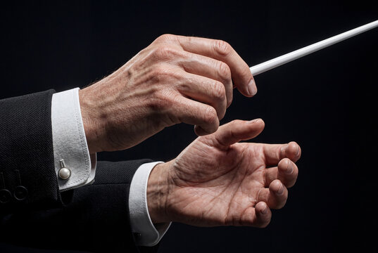 Close up of older masculine hands holding a white conductor baton against a dark setting