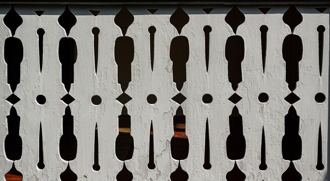 Close-up architectural shot of a vintage white wooden balcony railing featuring a traditional carved cutout motif and geometric repetition casting sharp shadows on the house floor.