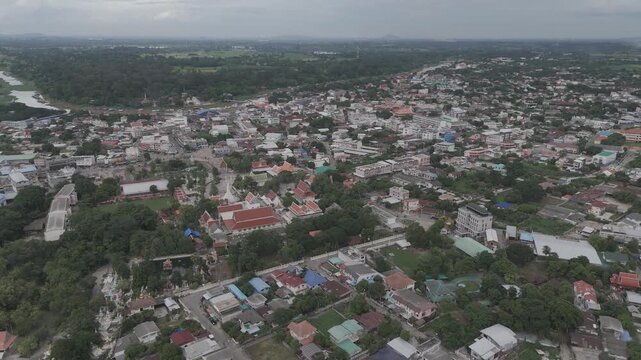 Aerial drone view of Uthai Thani city center in Thailand on an overcast day. Featuring local residential houses, streets, the Sakae Krang River, and a prominent Buddhist temple amidst lush green trees