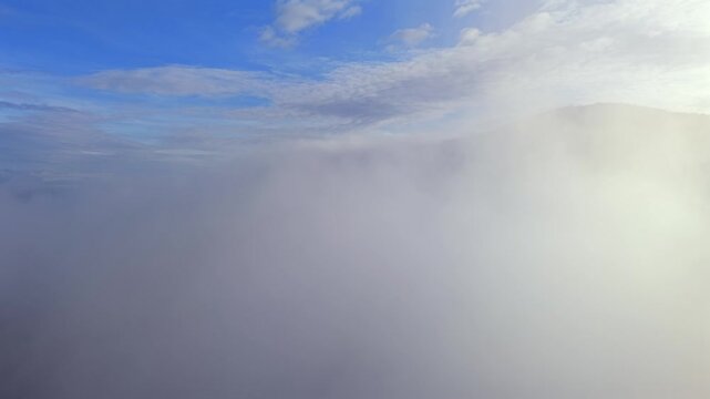 A cinematic aerial drone perspective capturing a thick, flowing blanket of morning clouds and mist sweeping gracefully across a mountain landscape. The scene features a bright, sunlit sky and the soft