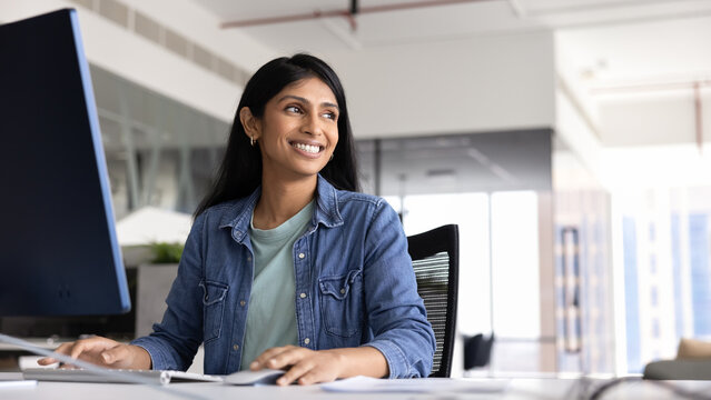 Cheerful Indian businesswoman sits at desk, working on pc, looks into distance with contented smile during workday in office. Workflow using modern device, internet resources, new AI digital tools