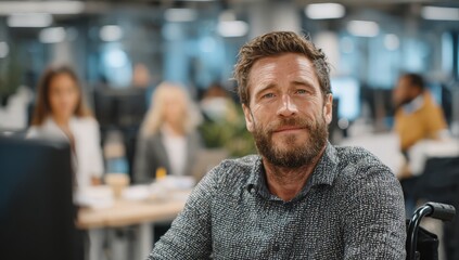 Obraz premium Male office worker with beard smiles at camera while seated in wheelchair, modern office environment with blurred colleagues in background