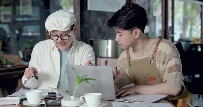 Asian father explains cafe management while son studies laptop, learning family restaurant operation, order planning, accounting paperwork, inventory control during practical mentorship lesson
