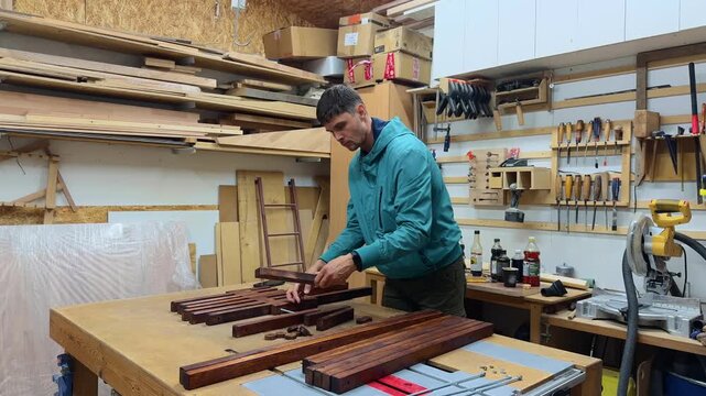 Skilled carpenter assembling wooden garden chair from stained slats on workbench inside home woodworking shop with organized hand tools, shelves, miter saw and authentic workshop lighting in wide shot