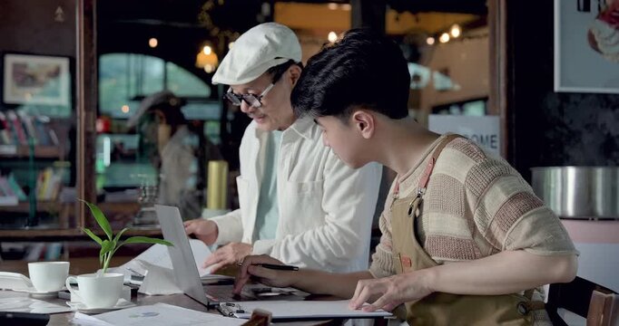 Asian father and son manage cafe documents with laptop, reviewing restaurant order list, supplier invoice, daily accounting, inventory update, family business handover planning together at work