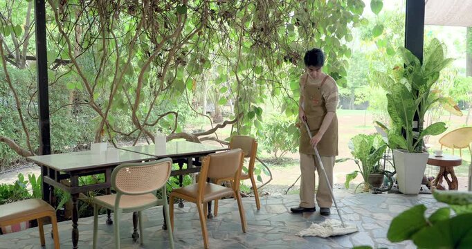 Asian son mops outdoor cafe dining area under leafy patio, following cleaning checklist, hygiene routine, table readiness, family restaurant training, service preparation, maintenance duty today.