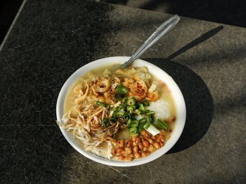 An image  of a bowl of traditional Indonesian chicken porridge or bubur ayam topped with shredded chicken, fried soybeans, chopped scallions, and cakwe slices on a stone table.