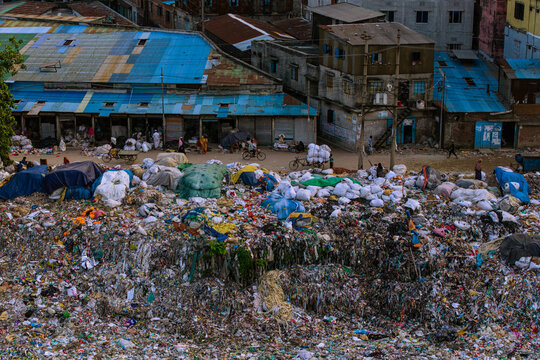 Aerial view of a massive garbage dump with piles of plastic waste and sacks near residential buildings with blue roofs in Keraniganj, Dhaka Division, Bangladesh.