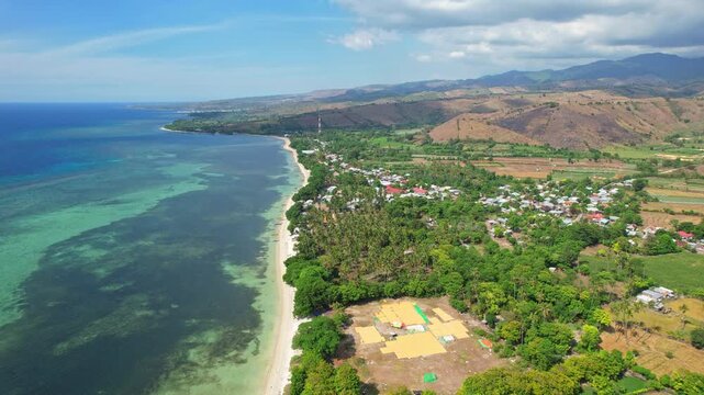 Aerial drone footage of a long white sand beach in Kilo city area, Bima regency, Sumbawa island, Indonesia, with blue crystal sea waters and reefs, village houses, and hills at the horizon