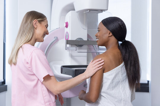 Female tech in pink scrubs guiding African-American woman undergoing mammography with clear paddle