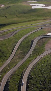 Vertical Screen: Beartooth Highway in Montana, USA captured from drone showing serpentine mountain road with cars navigating switchbacks through lush alpine meadows and snow patches.